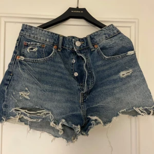 Blå jeansshorts från Zara - Snygga blå jeansshorts från Zara med slitna detaljer och fransar. De har en knappgylf och klassisk femficksdesign. Perfekta för en avslappnad stil.