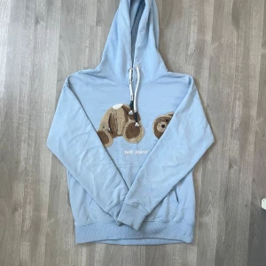 Ljusblå hoodie från Palm Angels - Säljer en ljusblå hoodie från Palm Angels med en unik nallebjörnsdesign på framsidan. Hoodien har en justerbar dragsko i huvan och är perfekt för en avslappnad stil. Den är både snygg och bekväm. Har en defekt på midjan