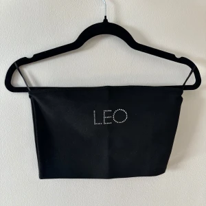 Kort bandeau topp - Svart kort bandeau topp med texten ”LEO” i strasstenar från NA-KD x Galore. Har silikon längst uptill på innersidan så den hålls bättre på plats. Använd 1 gång.