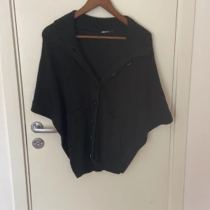 Svart stickad kofta från Gina Tricot - Säljer en svart stickad kofta från Gina Tricot med bred krage och stora knappar framtill. Koftan har korta ärmar och två praktiska fickor. Perfekt för lager-på-lager och chill stil. Passar dig som gillar loose fit och enkel design.