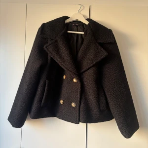 Svart teddyjacka från Chelsea Girls - Säljer en svart teddyjacka från Chelsea Girls med bred krage och dubbelknäppning med knappar. Jackan har en lite croppad passform och är tillverkad i ett mjukt, fluffigt material som ger en cool och mysig vibe.