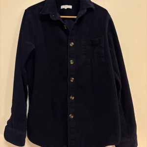 Mörkblå manchester overshirt S.C.W. - Snygg mörkblå overshirt från S.C.W. i mjuk manchester. Klassisk krage, bröstficka och knappar framtill. Långärmad och loose fit, perfekt att slänga över en t-shirt. Passar dig som gillar stilren och enkel design. Plagget har inga defekter och är perfekt inför hösten! Hör av dig vid frågor. (Plagget skickas inom 24h efter den köpts)