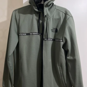 Grön vindjacka från The North Face - Snygg grön vindjacka från The North Face i storlek M. Jackan har ett coolt textur-mönster, huva med snörning och svarta detaljer med logga på bröstet och över bröstet. Perfekt för dig som gillar streetwear och outdoor-vibe.