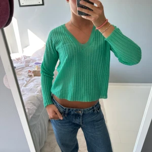 Grön ribbad stickad tröja med v-ringning - Snygg grön stickad tröja med ribbad struktur och v-ringad hals. Modellen är lite croppad och har långa ärmar. Perfekt att matcha med jeans för en chill och trendig look. Mjuk och skön, passar dig som gillar färg och enkel stil.