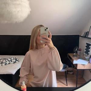 Ljusrosa långärmad klänning - Säljer en söt ljusrosa blus med långa puffärmar och rundad hals med lätt rynkning. Klänningen har en loose passform och är tillverkad i ett mjukt, slätt tyg som faller fint. Perfekt för dig som gillar enkel och stilren design.