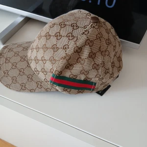 Gucci beige keps med GG-mönster - Snygg beige keps från Gucci med klassiskt GG-monogram över hela kepsen och en ikonisk grön-röd rand på sidan. Justerbar rem med läderdetalj baktill och svart insida med Gucci-logga. Perfekt för dig som gillar lyxiga accessoarer och vill sticka ut.
