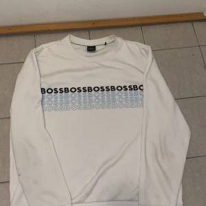 Vit långärmad tröja från BOSS bra för hösten - Vit långärmad tröja från BOSS med tryckt logga i svart och ljusblått över bröstet. Tröjan har rund hals och är gjord i mjuk bomull, perfekt för chill dagar. Enkel och clean design med ribbade muddar vid hals och ärmslut.