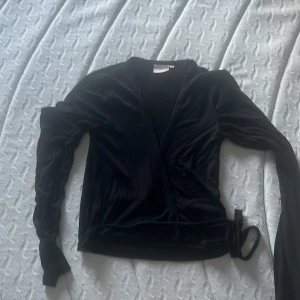 Svart omlottopp från Nelly - Svart omlottopp från Nelly i mjukt skönt tyg med långa ärmar och knyt i sidan. Snygg v-ringad design som ger en stilren look. Perfekt för dig som gillar trendiga och bekväma plagg med lite extra lyxig känsla.