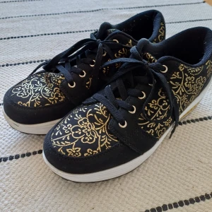 Svarta sneakers med guldfärgade blommor - Unika svarta sneakers från med guldfärgade blommönster.. Skorna har snörning, rund tå och en vit plattformssula med gul insats. Mjuka och lätta. Använda ca 5-7 gånger. 