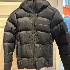 Svart jacka från Sail Racing - Säljer en svart jacka från Sail Racing i storlek S. Använd ca 3 gånger förra vintern. Perfekt för kalla dagar med sin fluffiga och varma design. Stilren look som funkar till det mesta.
