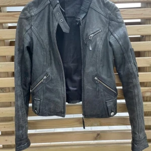 Svart skinnjacka med dragkedjor - Snygg svart skinnjacka med coola detaljer som dragkedjor på fickor och ärmslut, samt knappar vid midjan. Jackan har en figurnära passform och hög krage med spänne. Perfekt för dig som vill ha en edgy look.