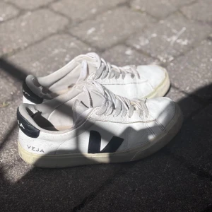 Vita Veja sneakers med svart V-logga - Säljer ett par Veja sneakers i vitt med svart V-logga på sidan och svart detalj vid hälen. Säljer biligt eftersom de är ganska slitna💗