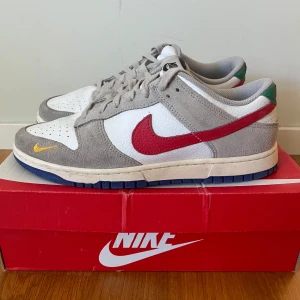 Nike Dunk Low vit/grå/röd EUR 44 - Nike Dunk Low sneakers med vit läderbas, grå mockadetaljer och röd Swoosh på sidorna. Yttersulan är mörkblå och det finns en liten gul Swoosh på tåpartiet. Skorna har snörning och en låg siluett, samt gröna detaljer vid hälen. Perfekt för dig som gillar streetwear.