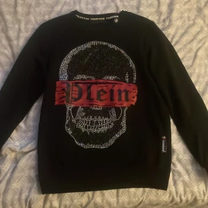 Svart tröja med döskalle från Philipp Plein - Svart långärmad tröja från Philipp Plein med stor döskalle framtill dekorerad med glittriga detaljer och röd banderoll med texten 'Plein'. Tröjan är gjord i 100% bomull och har en cool metall-logga i nacken. Perfekt för dig som gillar edgy streetwear.