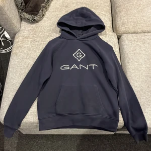 Mörkblå hoodie från GANT  - Snygg mörkblå hoodie från GANT med stort broderat logotyptryck på bröstet. Tröjan har huva, känguruficka och långa ärmar. Perfekt för chill dagar och enkel att matcha med jeans eller joggers. Materialet är mjukt och känns skönt mot huden.
