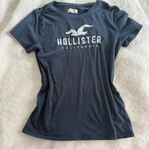 Mörkblå Hollister t-shirt med tryck - Snygg mörkblå t-shirt från Hollister med klassiskt vitt logotryck och fågel på bröstet. Modellen är figurnära med rund hals och korta ärmar. Mjuk och skön bomull som passar perfekt till jeans eller shorts. Enkelt och stilrent plagg för dig som gillar casual vibes.