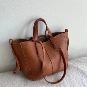 Brun handväska - Säljer en snygg brun handväska med två handtag och en lång axelrem. Väskan har en stilren design med struktur i materialet och är perfekt för att bära dina vardagsprylar. Enkel att matcha till olika outfits!