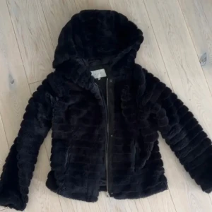 Svart pälsjacka med huva från Vila - Mjuk och fluffig svart pälsjacka i storlek S från Vila med huva och dragkedja framtill. Jackan har ett ribbat mönster i pälsen och är perfekt för dig som vill ha en varm och snygg look. 