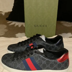 Gucci Ace  - Snygga svarta Gucci ace skor med klassiskt GG-mönster och vita sulor. Skorna har röda och blå ränder på sidan samt metallic-detaljer i rött och blått på hälen. Snörning framtill och rund tå. Perfekt för dig som vill sticka ut med en lyxig touch. 