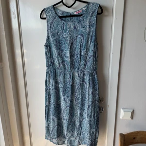 Blå paisleymönstrad klänning Indiska - Snygg blå paisleymönstrad klänning från Indiska i storlek M. Klänningen är ärmlös med rund halsringning och har en avslappnad passform med resår i midjan. Materialet känns lätt och luftigt, perfekt för varma dagar. Paisley-mönstret ger en bohemisk vibe.
