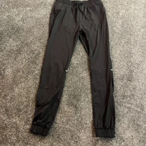 Svarta träningsbyxor från Famme - Svarta träningsbyxor från Famme i storlek M. Byxorna har elastisk midja med snörning, smal passform och reflexdetaljer runt knäna. Tillverkade i lätt syntetmaterial som andas, perfekta för löpning eller gym. Muddar vid benslut för extra komfort. Aldrig använts❤️