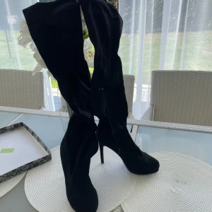Svarta höga stövletter med klack - Säljer ett par svarta, höga stövletter med smal klack och rund tå. Skorna är i mjuk mocka och har dragkedja på insidan för enkel på- och avtagning. Perfekta för dig som vill ha en edgy och stilren look.
