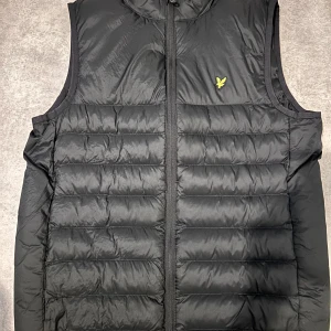 Svart dunväst från Lyle & Scott - Svart dunväst från Lyle & Scott med quiltad design och dragkedja framtill. Västen har ett litet gult logomärke på bröstet och är tillverkad i syntetmaterial. Perfekt för lager-på-lager och har en clean, sportig look.