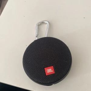 Svart rund JBL väska med karbinhake - Säljer en svart rund väska från JBL med karbinhake upptill, perfekt att fästa på väskan eller bältet. Väskan har ett grovt textilmaterial på framsidan och detaljer i plast. Den är liten och smidig, med ett diskret JBL-märke i rött framtill.