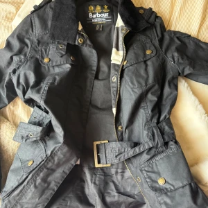 Svart Barbour parkajacka med bälte - Svart parkajacka från Barbour International med bälte i midjan, flera fickor med och dragkedja framtill. Jackan har krage i manchester och guldiga metalldetaljer. Perfekt för dig som gillar klassisk stil med edge.