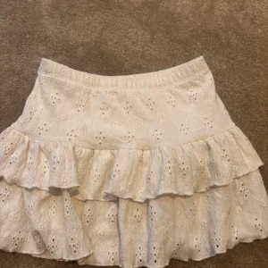 Vit volangkjol med broderade detaljer - Supersöt vit kortkjol med volanger och broderade hålmönster. Kjolen har en elastisk midja och är gjord i mjukt bomullstyg. 