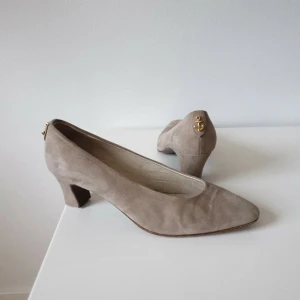 Beige mocka pumps med gulddetalj - Snygga beige pumps från Rizzo i mjuk mocka med spetsig tå och låg klack (behöver klackas om). På hälen sitter en cool guldfärgad ankardekor som ger extra stilpoäng. Perfekta för dig som gillar klassisk design med en twist. Tillverkade i Italien.