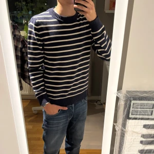 Marinblå randig stickad tröja - Säljer en marinblå stickad tröja med vita horisontella ränder. Tröjan har rund halsringning, ribbade muddar vid ärmslut och nederkant samt långärmad passform. Perfekt för dig som gillar klassisk och chill stil.