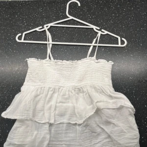 Vit kort sommarklänning med volang - Supersöt vit klänning med smala axelband och volangdetaljer. Överdelen är smockad och klänningen har en luftig, lätt känsla som passar perfekt för varma dagar. Stilren och enkel design som är lätt att matcha med accessoarer.