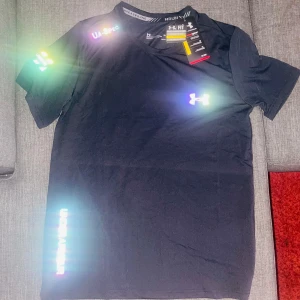 Svart Under Armour tränings-tshirt - Svart t-shirt från Under Armour med tryck på bröstet och ärmen, samt texten 'UNDER ARMOUR' längs sidan. Modellen är kortärmad och har rund hals. Materialet är lätt och andas, perfekt för träning. Snygg, sportig look med slim passform.