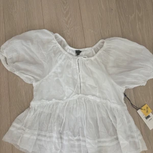 Vit blus med puffärm från Amelie & E - Supersöt vit blus från Amelie & E i storlek S. Blusen har puffiga korta ärmar, volang nedtill och en söt knytning vid halsen. Tillverkad i ett lätt och luftigt bomullstyg som ger en romantisk vibe. Perfekt för dig som gillar en feminin och trendig stil.