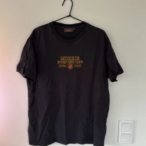 Svart Morris t-shirt med tryck - Svart t-shirt från Morris Sporting Club med gul text och klubbmärke tryckt på bröstet. Klassisk rund hals och korta ärmar. Skön bomullskvalitet och normal passform. Perfekt för dig som gillar stilrena och sportiga plagg med en touch av tradition.