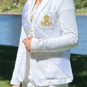 Vit kavaj med gulddetaljer RL - Snygg vit kavaj från Ralph Lauren med guldfärgade knappar och ett broderat RL-emblem i guld på bröstfickan. Kavajen har klassisk passform, två fickor fram och är tillverkad i ett mjukt bomullsmaterial. Perfekt för student eller andra speciella tillfällen. Har en liten fläck som märka it så tydligt 