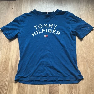Blå t-shirt Tommy Hilfiger - Snygg blå t-shirt från Tommy Hilfiger med vit logotyp och flagga tryckt på bröstet. Klassisk rund hals och korta ärmar. Perfekt för dig som gillar stilrena märkesplagg och vill ha en enkel, fräsch look. Materialet är mjuk bomull som känns skönt mot huden.