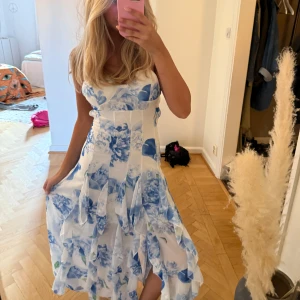 Blommig långklänning med volanger - Superfin långklänning från Bella barnett med blåa och vita blommor, omlottvolanger och smala axelband. Klänningen har en öppen rygg med snygg cut-out och sitter bekvämt med figurnära passform. (Finns ej kvar)