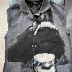 Svart väst med vargmotiv från Novamen - Unik svart väst från Novamen med stort vargtryck över hela framsidan och baksidan. Västen har krage, tryckknappar och är tillverkad i ett grovt, vävt material. Perfekt för dig som vill sticka ut med en edgy och konstnärlig look.