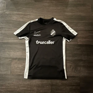 Svart AIK fotbollströja Nike - AIK fotbollströja i svart med vita paneler längs sidor och axlar. Märkt med AIK:s klubbmärke på bröstet, Nike-logga och sponsorer som Truecaller, Stadium, Fabege och SVEA. Tröjan är kortärmad och tillverkad i lätt, ventilerande polyester. Låsnat lite färg på aik logan men knappt märkbart