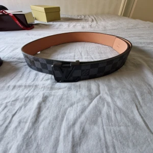 Svart rutig Louis Vuitton skärp i skinn - Säljer ett stilrent Louis Vuitton bälte i svart och grått rutigt mönster med klassisk LV-logga på spännet. Bältet är tillverkat i äkta skinn med ljusbrun insida och har en bred, platt form. Perfekt för dig som vill ha en lyxig detalj till outfiten.