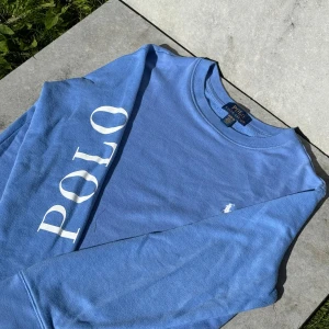 Blå sweatshirt från Polo Ralph Lauren - Snygg blå sweatshirt från Polo Ralph Lauren med stort vitt POLO-tryck längs ena ärmen och klassisk broderad logga på bröstet. Rund hals och långa ärmar, tillverkad i mjuk bomull. Perfekt för dig som gillar stilrena och sportiga vibes.