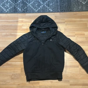 Svart quiltad cardigan Polo Ralph Lauren - Svart cardigan från Polo Ralph Lauren i storlek S. Jackan har quiltade ärmar och huva, dragkedja framtill och två fickor. Materialet är polyester och syntet, den enda skadan är nere på ärmen. Perfekt nu till hösten!
