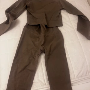Bruna ribbade set -  Ett tränings sett i ribbat tyg, bestående av en långärmad topp och ett par byxor. Setet är i en neutral brun/grå-beige ton. Toppen har rund halsringning och är något kortare i modellen (croppad), medan byxorna är höga i midjan med en smal passform.   Från: Divina Storlek: S, både över och under delen Använd en gång max 2 