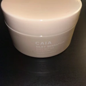 CAIA Silky Sensation Body Butter - Lyxig body butter från CAIA i en stilren beige burk. Konsistensen är krämig och vit, perfekt för att återfukta huden. Innehåller sheasmör och macadamiaolja som ger en mjuk och silkeslen känsla. Passar alla hudtyper och har en fräsch doft.