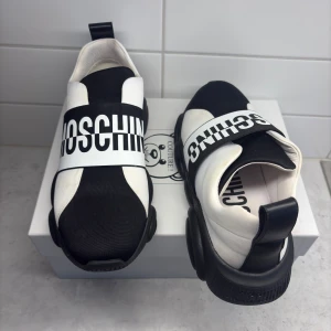 Moschino svartvita sneakers strl 40 - Säljer ett par Moschino sneakers i svart och vitt med bred logorem över foten. Sulan har en unik design med nallemotiv och texten 'This is not a Moschino toy'. Ovandelen är i textil och syntet, med rund tå och platt sula. Perfekt för dig som gillar statement pieces. Använda sparsamt fåtal gånger. Säljs inte i dessa färger längre och är slutsålda. Intresse koll.