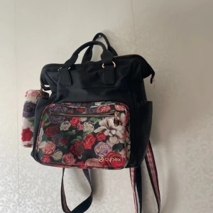 Blommig ryggsäck från Cybex - Säljer en svart ryggsäck från Cybex med blommigt mönster i rött, lila, beige och grönt på både utsidan och insidan. Väskan har guldfärgade detaljer, dragkedja och flera fack. Axelremmarna är randiga och väskan har både handtag och axelremmar.