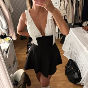 Zara klänning  - Säljer en snygg svartvit klänning med djup v-ringning och breda axelband. Kjolen är svart och har en utsvängd form, medan överdelen är vit och har en omlott-look. Klänningen är ärmlös och har en modern, chic vibe som passar perfekt för dig som gillar att sticka ut. Aldrig använd 