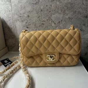 Ny beige quiltad axelväska - Supersnygg beige axelväska med klassiskt quiltat mönster och guldfärgad kedja. Väskan har ikonisk CC-logga framtill, praktiskt innerfack med dragkedja och lyxig röd insida. Perfekt storlek för det viktigaste och en riktig statement piece. Oanvänd med plast och etikett på. 